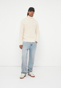 Maglione a collo alto lavorato a maglia crema, jeans dritti azzurri chiari e sneakers beige con suole marroni; un cappellino nero completa l'outfit.