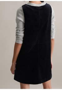 Femme portant une robe sans manches en velours côtelé noir sur un t-shirt gris à manches longues côtelé, vue de dos sur un fond uni.