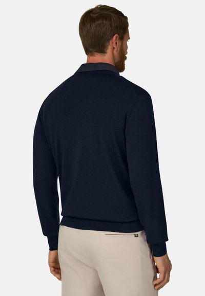 Homme aux cheveux courts et bruns avec une barbe, portant un pull bleu marine foncé et un pantalon beige clair, vu de dos sur un fond uni.