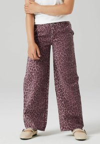 Bērns, kas valkā plašas purpura leopardu apdrukas bikses, baltu topu un krēmkrāsas studētus kurpes, stāv ar rokām sastieptām.