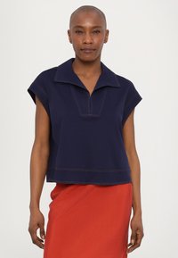Navy blue cropped polo top with a pointed collar and short sleeves, featuring contrasting beige stitching and a quarter zip at the front.