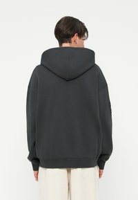 Σκούρο γκρι φούτερ με μεγάλη κουκούλα, χαλαρή εφαρμογή, πεσμένα ώμους και ριμπ μανσέτες. Λείο βαμβακερό ύφασμα, απλός σχεδιασμός, χωρίς ορατά μοτίβα.