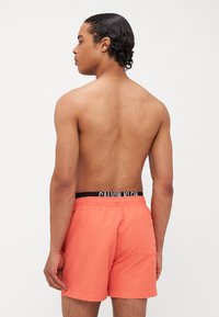Calvin Klein svømmeshorts i korall med en elastisk svart midje strikk med logo. Jevnt, lett stoff, med en baklomme detalj.