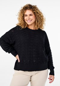 Pull en maille noir avec un motif texturé, des poignets côtelés et un ourlet festonné. Associé à un pantalon beige, il offre une coupe décontractée.