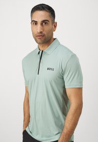 Vaalea vihreä polo-paita, jossa on musta vetoketjusulkiminen ja tekstuurinen kuvio, sekä musta "BOSS"-logo rinnassa. Lyhyet hihat.