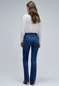 Jeans acampanados de un azul oscuro con una cintura de media altura, combinados con una blusa blanca de manga larga. Presentan una textura suave y bolsillos traseros visibles.