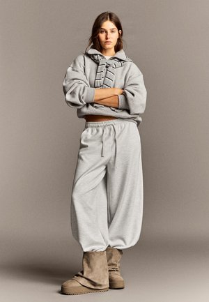 Jeune femme debout, les bras croisés, portant un sweat à capuche gris oversized, un pantalon de jogging gris clair ample et des bottes en daim beige sur un fond uni.