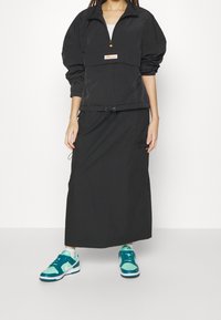 Czarna kurtka przeciwwiatrowa z półzamkiem, z oversize'owymi rękawami i regulowanym dołem, zestawiona z czarną maxi spódnicą i turkusowymi sneakersami.