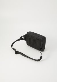 Sac à bandoulière rectangulaire noir avec un design texturé à pois sur un côté, une sangle ajustable et un tissu lisse de l'autre côté.