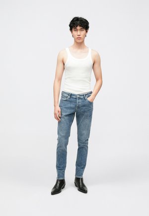 Débardeur blanc, jean bleu clair et bottines noires. Le jean présente un design classique à cinq poches. Texture lisse, détails minimaux.