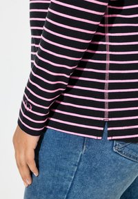 Černý top s dlouhým rukávem s růžovými pruhy, s bočním rozparkem a prošitými detaily. Spárován s denimem, zobrazující detail rukávu zblízka.