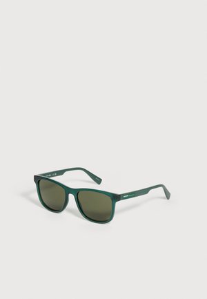 Lunettes de soleil rectangulaires vert foncé avec verres teintés et logo Lacoste sur la branche, placées sur un fond clair.