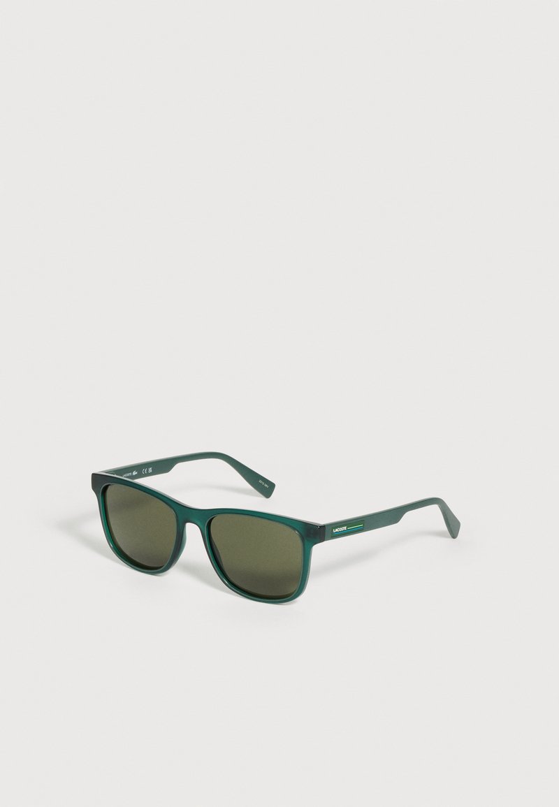 Lunettes de soleil rectangulaires vert foncé avec verres teintés et logo Lacoste sur la branche, placées sur un fond clair.