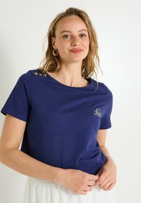Camiseta azul marino hecha de tela suave, con escote redondo, mangas cortas y botones dorados decorativos en el hombro. Incluye bordado de "Love".