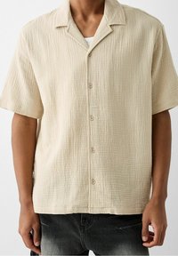 Beige kortärmad skjorta av texturerad tyg, med knapprad framtill och en camp collar-design. Subtil rutmönster synligt.