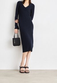 Robe tricotée navy avec un décolleté en V, des manches longues et une silhouette ajustée. Assortie à des sandales à talons noires avec des lanières et un petit sac à main noir.