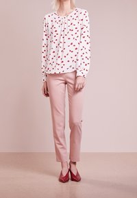 Femme portant un chemisier blanc à manches longues avec des motifs de feuilles rouges, un pantalon slim rose et des chaussures rouges à bout pointu, debout devant un fond uni.