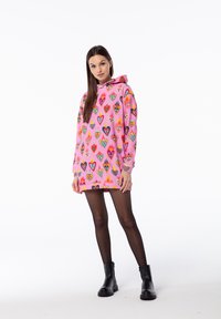 Rosa Kapuzenpullover mit bunten Herzmustern und Flammen. Das Model kombiniert ihn mit schwarzen Leggings und Ankle Boots. Weiche Textur.