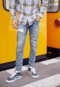 Camisa de cuadros azul claro con botones, vaqueros azules desgastados y deportivas verdes con detalles blancos, sobre un fondo amarillo brillante.