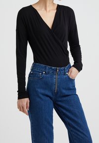 Schwarzer langärmliger Body mit V-Ausschnitt und Wickeldesign, kombiniert mit hoch taillierten blauen Jeans aus Denim, die einen Reißverschluss und Fronttaschen haben.