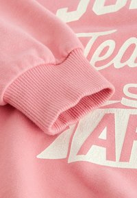 Rosa sweatshirt med ribbade ärmar och en mjuk textur. Har vit text med en spruckna yta. Närbild som framhäver tygdetaljer.