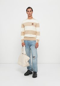 Beige och vit randig stickad tröja med rund halsringning och logoplagg, tillsammans med ljusblå ripped jeans och en krämfärgad tote-väska. Svarta sneakers.