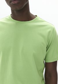 Camiseta de algodón verde claro con cuello redondo, mangas cortas y textura suave. Sin patrones visibles ni detalles de hardware.