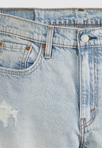 Jean en denim bleu clair avec bouton frontal, passants pour ceinture, poche avant et une petite déchirure près de la cuisse gauche.