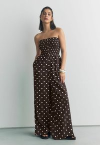 Salopetă maro, fără bretele, cu buline albe, având un corset elastic, nasturi în față și buzunare laterale. Material neted și picioare largi.
