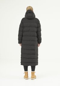Manteau long noir en duvet avec capuche, présentant des sections matelassées horizontales, des fentes sur les côtés et une texture lisse et brillante. Assorti à des bottes beiges.