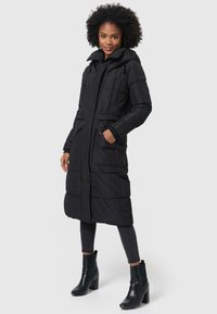Abrigo largo de plumas negro con capucha, diseño acolchado, bolsillos laterales y puños acanalados en contraste. Lleva puesto sobre unos jeans oscuros y botas de tacón.