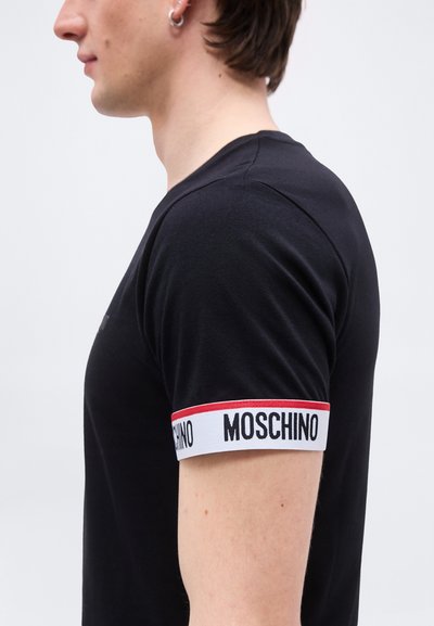 Jeune homme portant une chemise noire à manches courtes avec un bord blanc et rouge sur la manche, ornée du texte "MOSCHINO" en lettres noires.