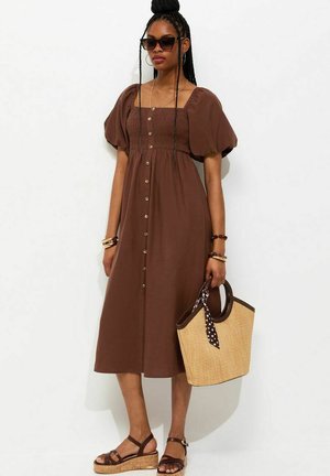 Femme portant une robe midi marron boutonnée avec des manches bouffantes, des lunettes de soleil foncées, des sandales, des bracelets, et tenant un sac en paille avec un foulard à pois.