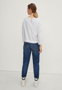 Top bianco a maniche lunghe con scollatura quadrata e dettaglio a chiave sul retro, abbinato a jeans in denim blu e sneaker bianche.