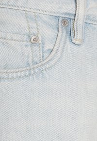 Světle modré džíny mají detailní pohled na pás, který zvýrazňuje stříbrné kovové prvky a kontrastní švy podél okrajů.