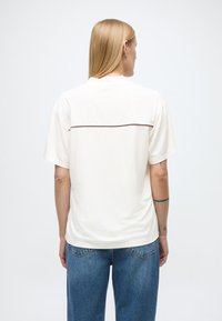 Chemise blanche à manches courtes avec un col rond et un motif de couture horizontale sur le haut du dos, associée à un jean bleu à jambes larges.