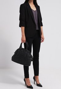 Sort vævet håndtaske med dobbelte håndtag, struktureret overflade og metalornamenter. Båret med en sort blazer, grå top og sorte bukser.