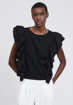 Ung kvinde med kort hår iført en sort ærmeløs top med flæset hulblonde og hvide bukser med højtaljede mod en ensfarvet baggrund.