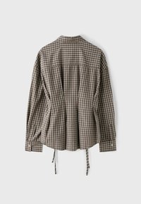 Chemise à carreaux marron et blanc à manches longues avec poignets boutonnés et liens latéraux ajustables, vue de dos sur un fond uni.