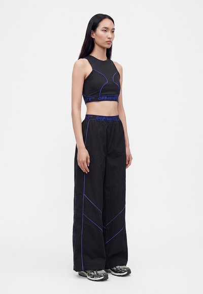 Γυναίκα που φοράει μαύρο crop top και φαρδιά παντελόνια με μπλε επένδυση και ζώνες μέσης με την επωνυμία "Off-White", στέκεται μπροστά σε λευκό φόντο.