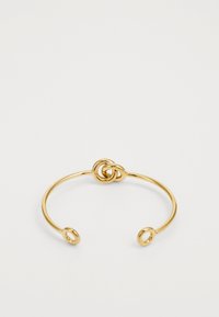 Gull armbånd i gull med en knutelignende design i midten, åpne ender med sirkulære detaljer, glatt metalloverflate. Minimalistisk stil.