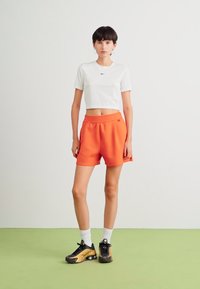 Baltas trumpas marškinėlis su juodu logo, oranžinės šortai su šoninėmis kišenėmis ir juodi sportbačiai su gelsvais akcentais. Modelis stovi ant žalios grindų.