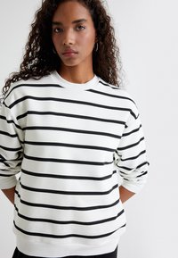 Sudadera blanca con rayas horizontales negras, cuello redondo y mangas largas. Textura suave con dobladillo y puños acanalados.