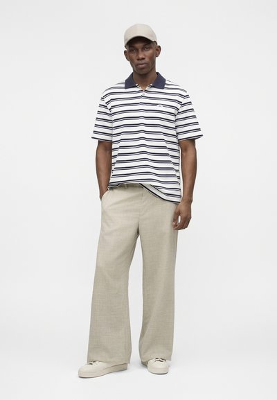 Άνδρας με μπεζ καπέλο, λευκό και ναυτικό ριγέ πόλο μπλουζάκι, μπεζ παντελόνια με φαρδιά μπατζάκια και λευκά αθλητικά, στέκεται μπροστά σε ένα απλό φωτεινό φόντο.