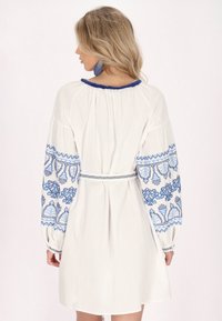 Witte jurk met blauwe geborduurde bloemenpatronen op lange mouwen, met een gathered halslijn, een taille met ceintuur en een soepele katoentextuur.