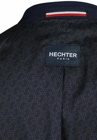 Tissu noir avec un motif géométrique bleu subtil. Comprend une étiquette indiquant "HECHTER PARIS" et un accent rouge, blanc et bleu au niveau de l'encolure.