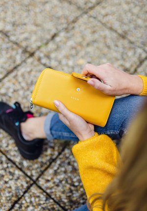 Gelber Reißverschluss-Wallet mit strukturierter Oberfläche, ausgestattet mit einem sichtbaren Markenlogo "VUCH" und einem Druckknopfverschluss. Eine Hand hält das Wallet.