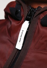 Maroonfärgad tygjacka med en svart dragkedja och en vit G-Star Raw-tag. Funktioner inkluderar en strukturerad yta och kontrasterande sömmar.