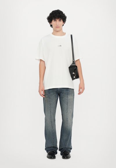 T-shirt blanche oversize avec un petit imprimé, associée à un jean bleu délavé. Sac à bandoulière noir avec détail à pression, chaussures noires sur un fond uni.