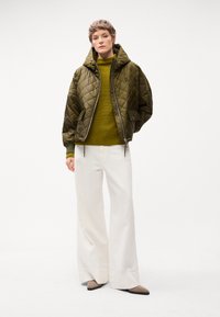 Gepolsterte olivgrüne Bomberjacke mit Kapuze, kombiniert mit einem gerippten olivfarbenen Pullover, weißen weit geschnittenen Hosen und beigen spitzen Ballerinas.